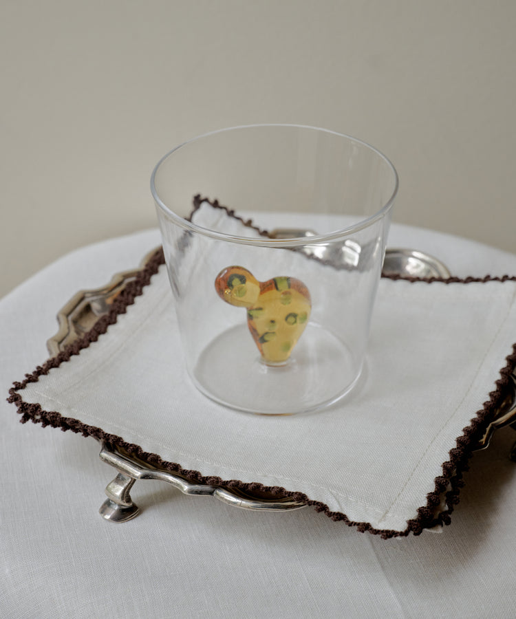 An Agave Tumbler from the set of 4 holds a small, multicolored abstract object. The glass sits on a white napkin with brown trim, neatly arranged on a silver tray.