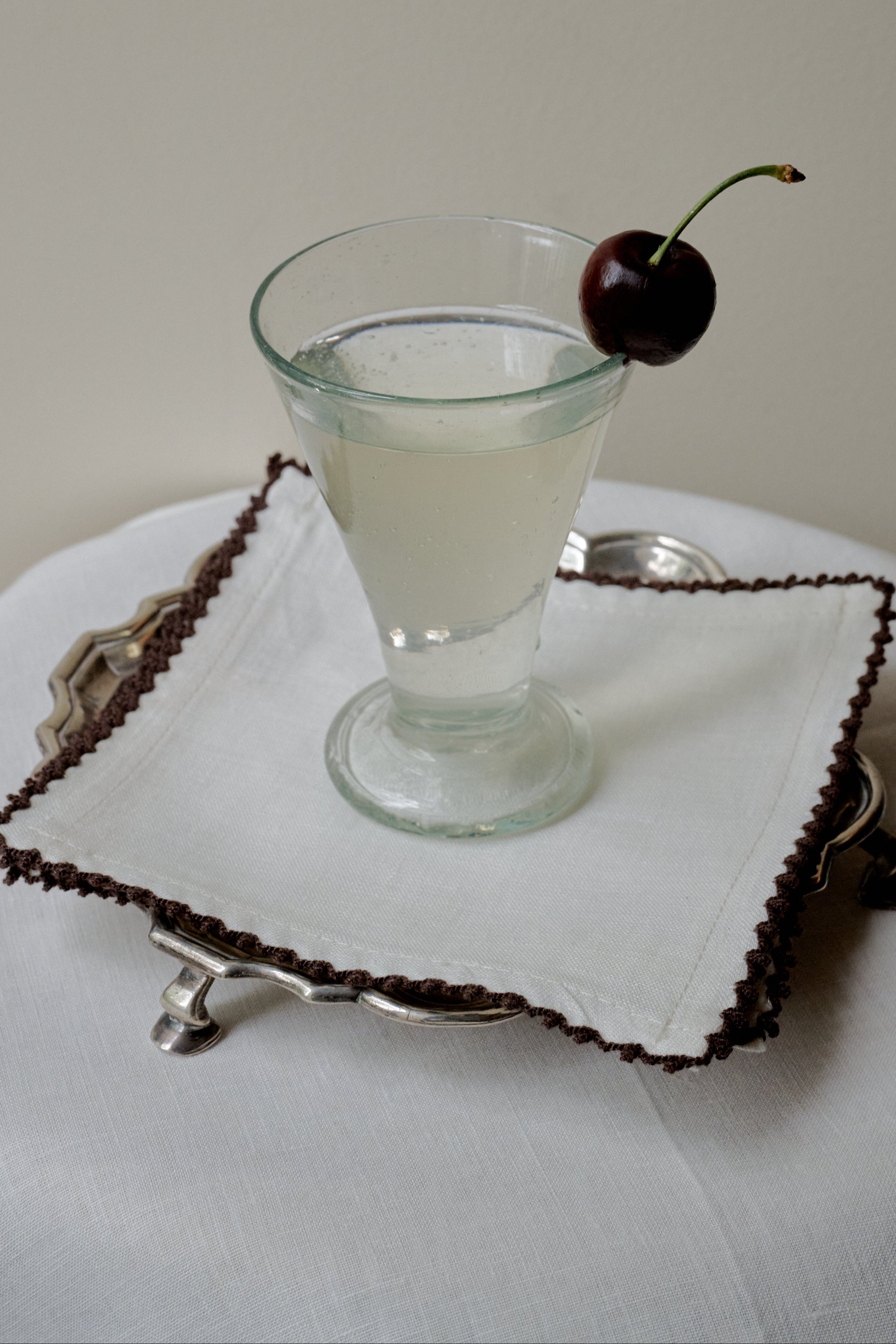 A clear drink in a stemmed glass, garnished with a dark cherry, sits atop the Nico Cocktail Napkin, Set of 4, placed on a silver tray over a white tablecloth.