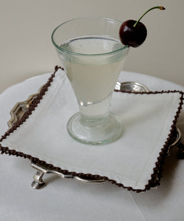 A clear drink in a stemmed glass, garnished with a dark cherry, sits atop the Nico Cocktail Napkin, Set of 4, placed on a silver tray over a white tablecloth.