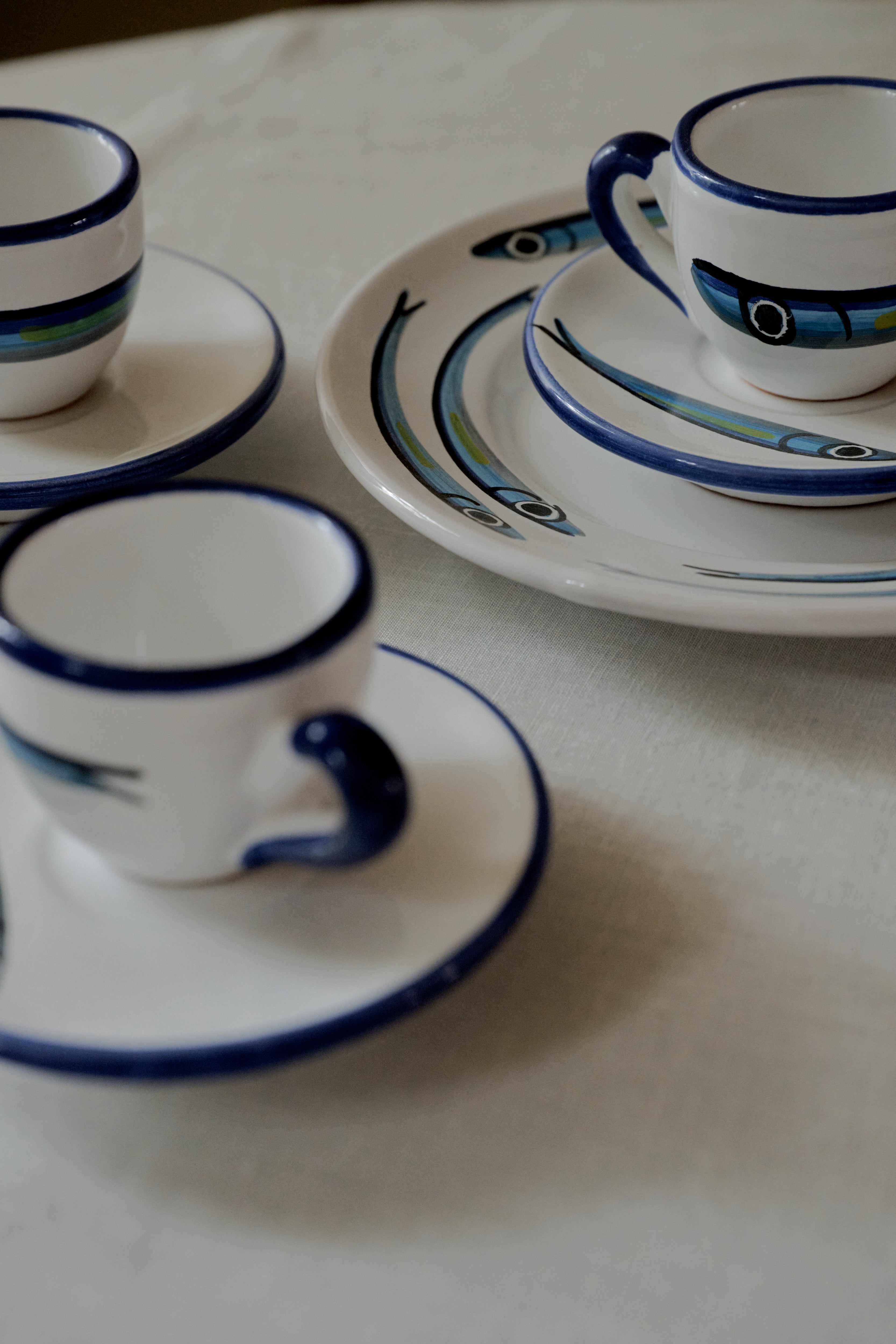 The Tommaso Espresso Set includes three espresso cups with saucers and two plates, all adorned with blue rims and fish designs, capturing a charming Mediterranean vibe on a white tablecloth.
