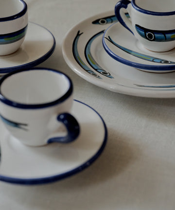 The Tommaso Espresso Set includes three espresso cups with saucers and two plates, all adorned with blue rims and fish designs, capturing a charming Mediterranean vibe on a white tablecloth.