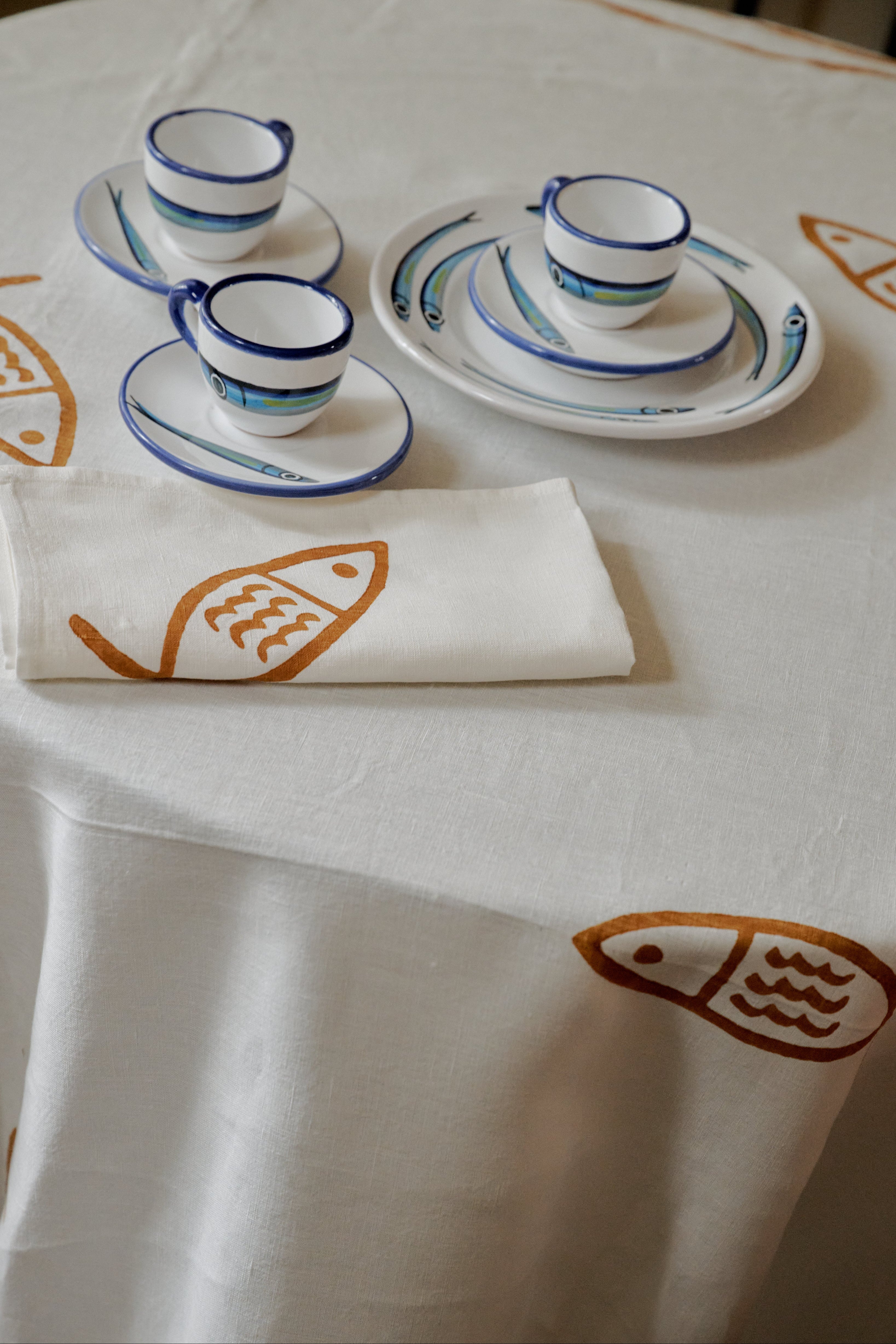 The Pezza Tablecloth, featuring white linen adorned with orange fish prints, is paired with two cups, saucers, and plates decorated with blue fish motifs, as well as a folded white napkin—an ideal choice for bringing a coastal touch to your dining experience.