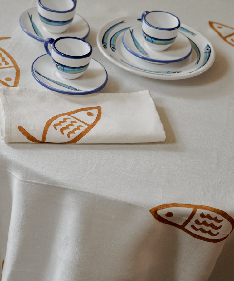 The Pezza Tablecloth, featuring white linen adorned with orange fish prints, is paired with two cups, saucers, and plates decorated with blue fish motifs, as well as a folded white napkin—an ideal choice for bringing a coastal touch to your dining experience.
