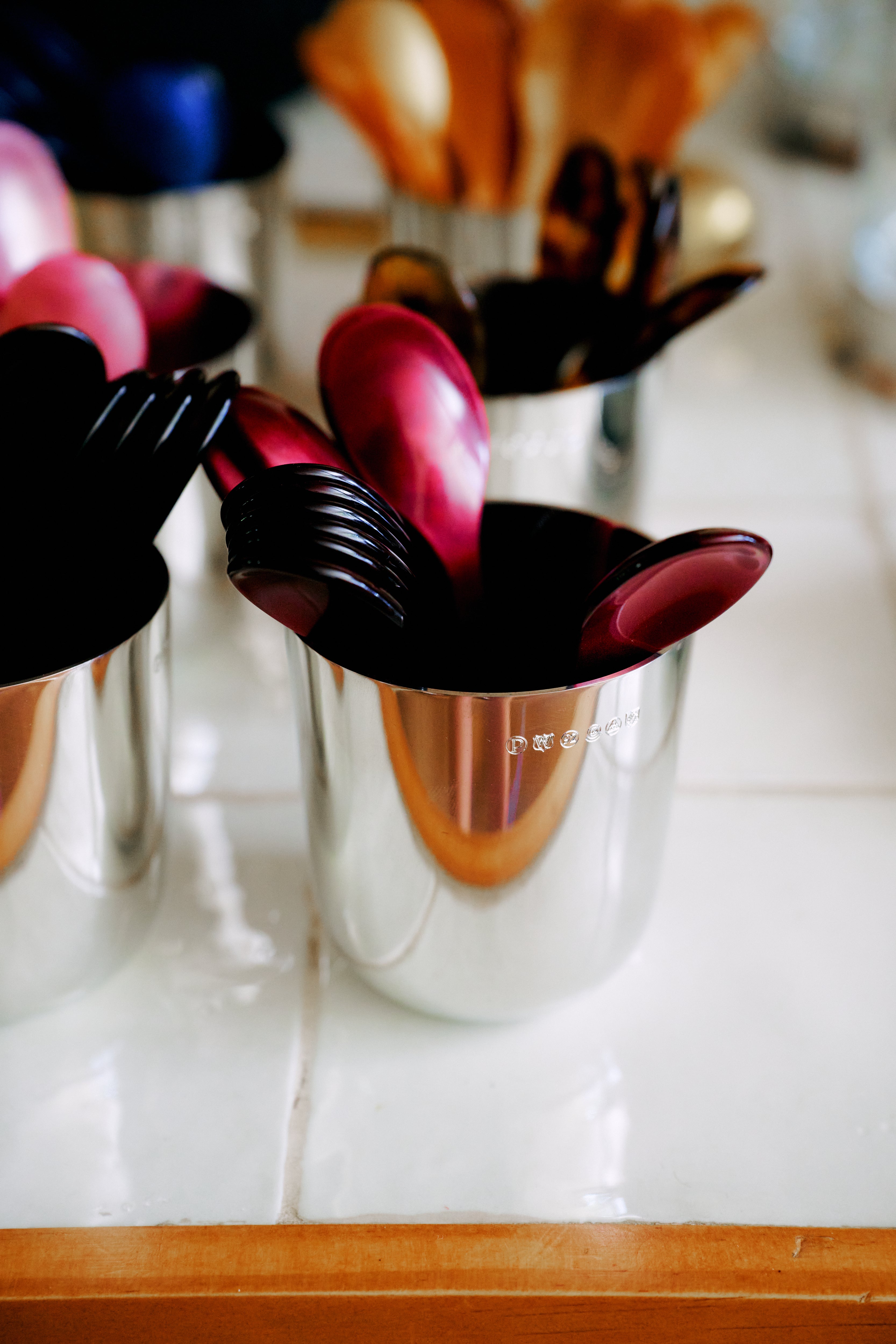 A metal cup holds glossy red and black Piccolo Spoons and forks, arranged on a white tiled surface with additional utensils in the background. These dishwasher-safe Piccolo Spoons are perfect for everyday use and bring a vibrant touch to your table setting.