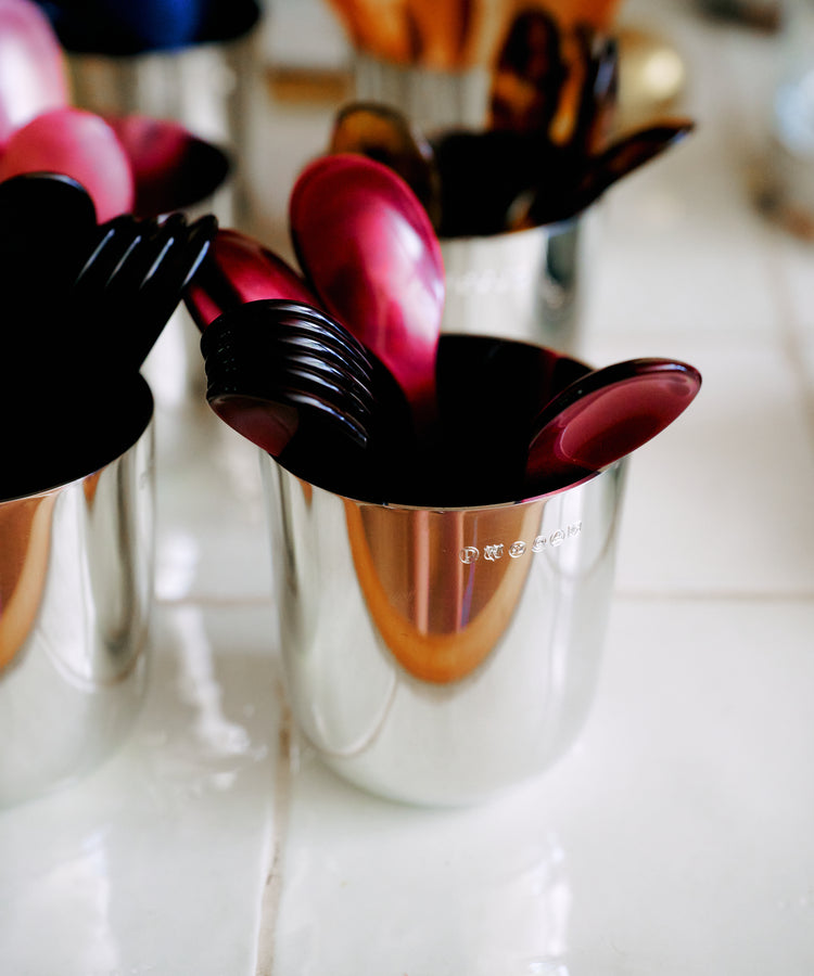A metal cup holds glossy red and black Piccolo Spoons and forks, arranged on a white tiled surface with additional utensils in the background. These dishwasher-safe Piccolo Spoons are perfect for everyday use and bring a vibrant touch to your table setting.