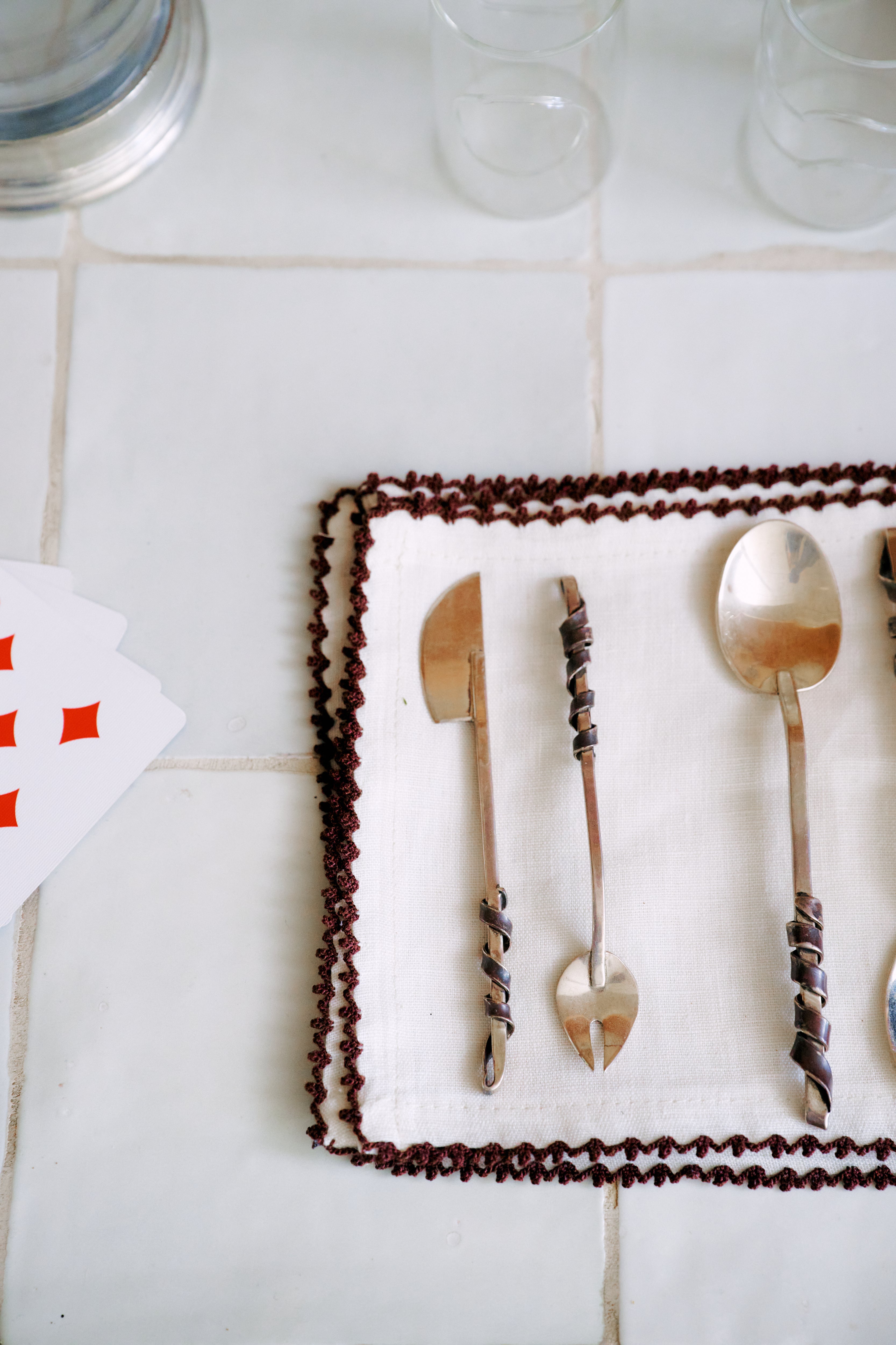 Three ornate metal utensils from the Heath Wagoner | Aperitivo Set—jewelry for your table—rest on a white cloth with a brown border, beside two empty glasses and partially visible playing cards atop a tiled surface, ready for your next aperitivo or evening snack.