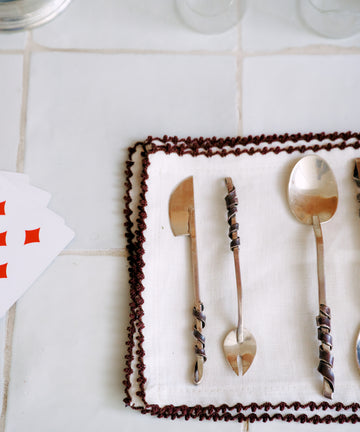 Three ornate metal utensils from the Heath Wagoner | Aperitivo Set—jewelry for your table—rest on a white cloth with a brown border, beside two empty glasses and partially visible playing cards atop a tiled surface, ready for your next aperitivo or evening snack.