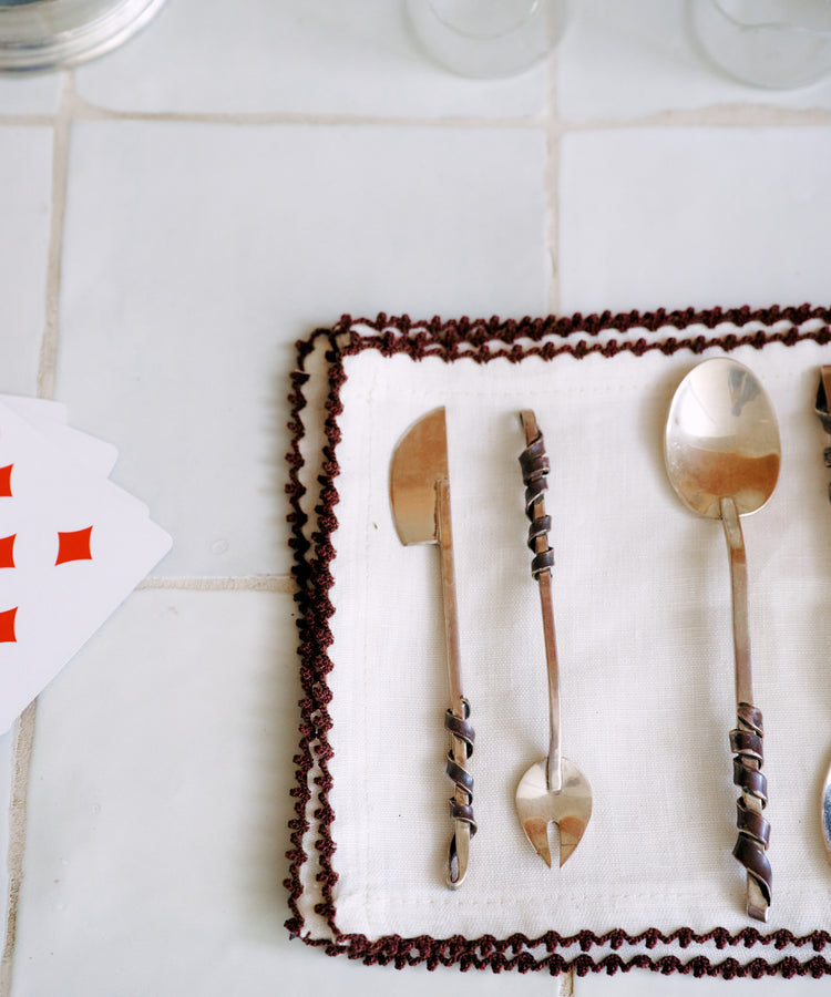 Three ornate metal utensils from the Heath Wagoner | Aperitivo Set—jewelry for your table—rest on a white cloth with a brown border, beside two empty glasses and partially visible playing cards atop a tiled surface, ready for your next aperitivo or evening snack.