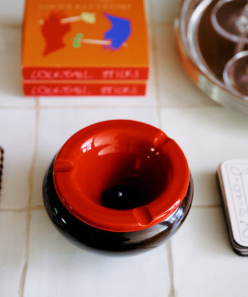 The Mix & Match Ashtray, a chunky ceramic piece handmade in Italy, rests on a tiled surface beside a stack of books and a silver tray.