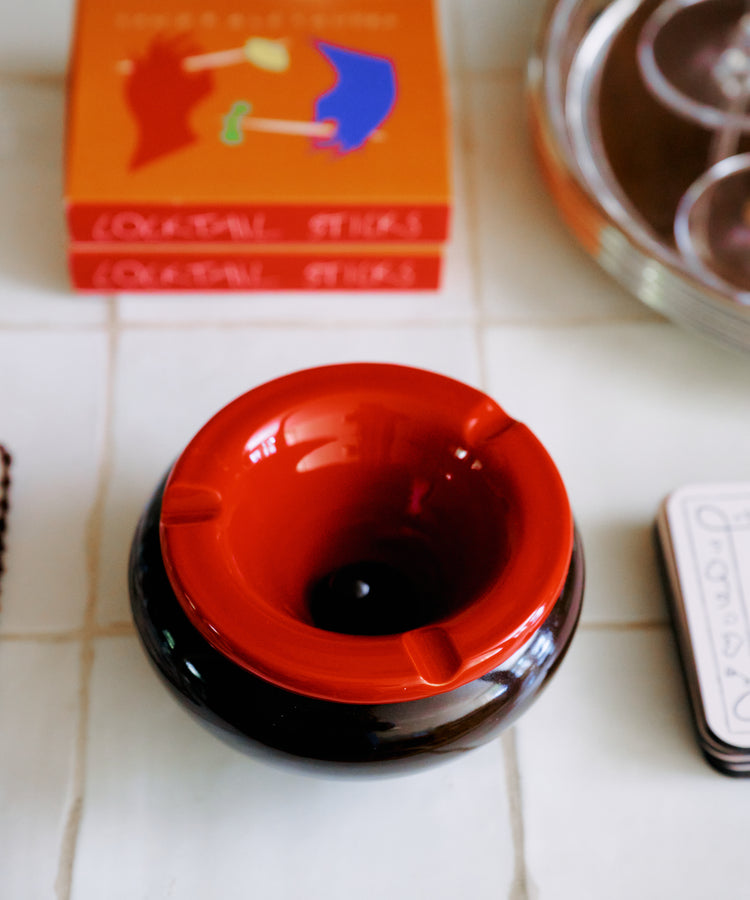 The Mix & Match Ashtray, a chunky ceramic piece handmade in Italy, rests on a tiled surface beside a stack of books and a silver tray.