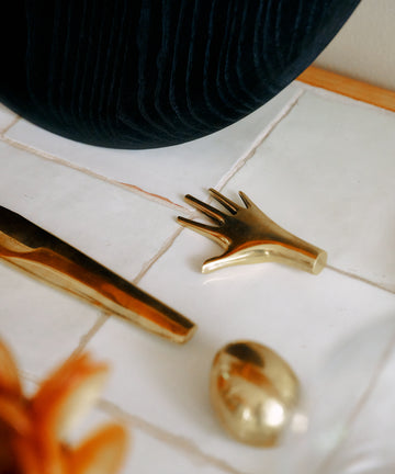 A Hand Paperweight in solid brass—evocative of Viennese modernism—rests alongside other brass forms on a white tiled surface near a dark textured fabric. Crafted by the Auböck workshop, these pieces exemplify timeless craftsmanship.