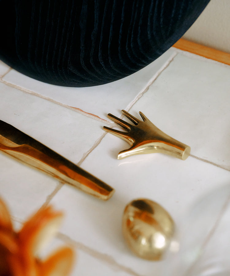 A Hand Paperweight in solid brass—evocative of Viennese modernism—rests alongside other brass forms on a white tiled surface near a dark textured fabric. Crafted by the Auböck workshop, these pieces exemplify timeless craftsmanship.