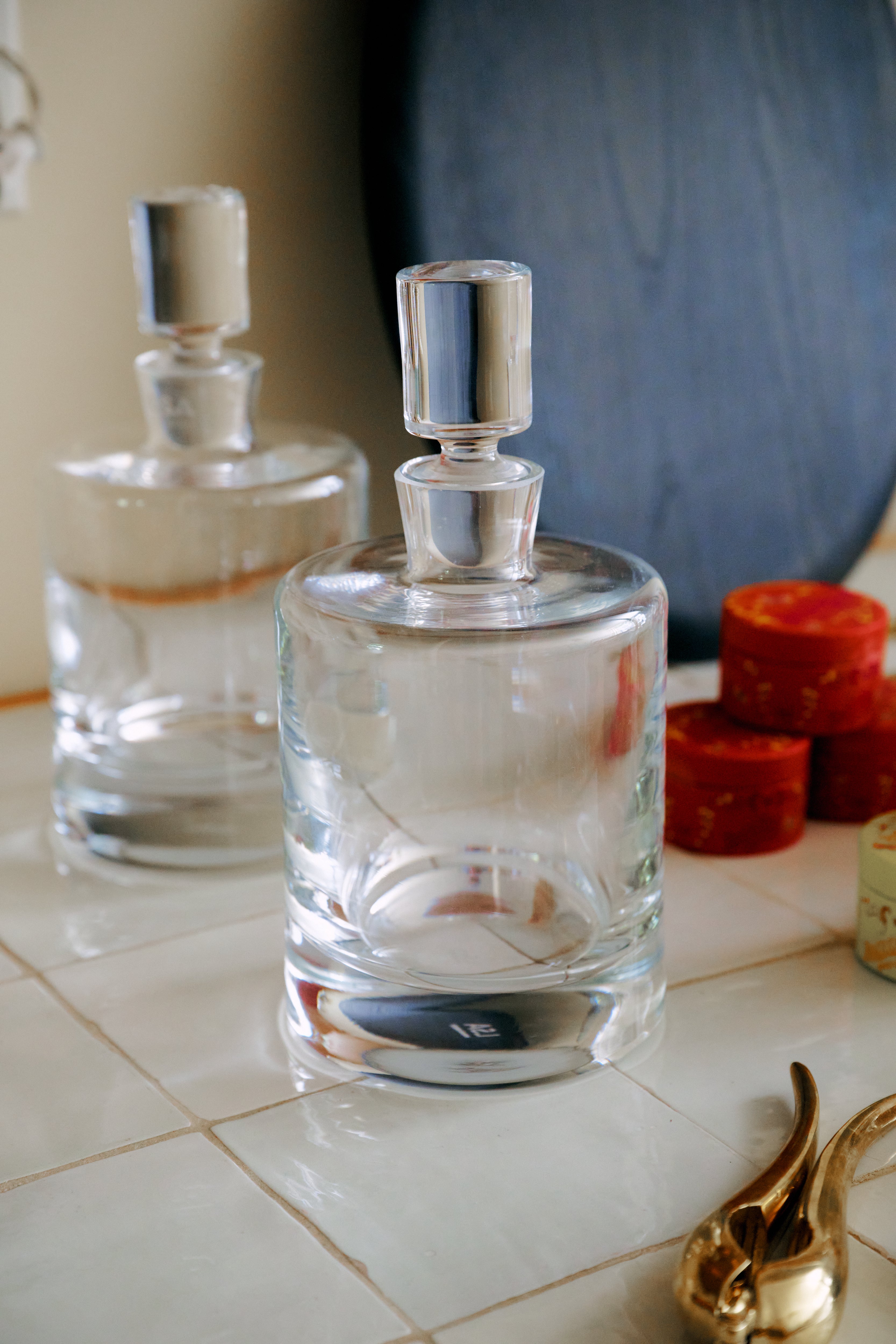 Two Boris Decanters with cylindrical stoppers sit on a tiled surface beside several small red boxes and a curved gold object, their elegant architectural profile adding a refined touch to the scene.