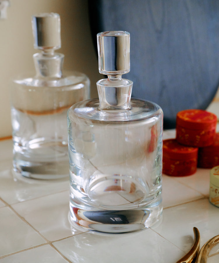Two Boris Decanters with cylindrical stoppers sit on a tiled surface beside several small red boxes and a curved gold object, their elegant architectural profile adding a refined touch to the scene.