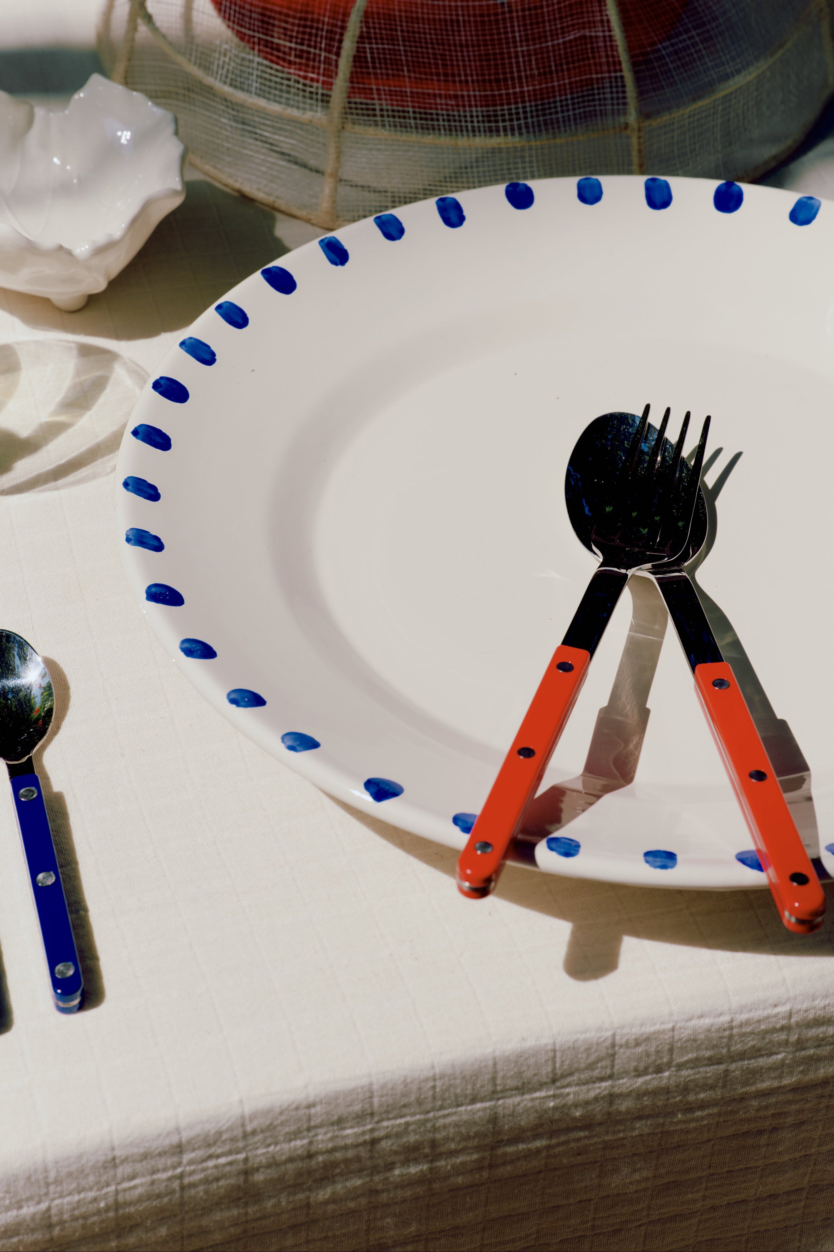 A white ceramic plate with a blue polka-dot rim sits on a tablecloth, styled with matching cutlery featuring bright orange handles and black metal heads.