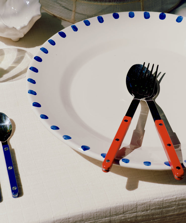 A white ceramic plate with a blue polka-dot rim sits on a tablecloth, styled with matching cutlery featuring bright orange handles and black metal heads.