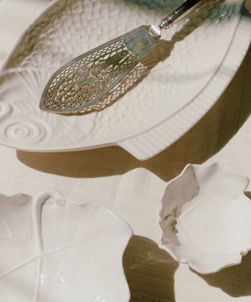The Peixe Platter, crafted from white ceramic in a sculptural leaf design, serves as a charming focal point alongside a decorative metal pie server, all arranged on a light-colored tablecloth. Sunlight casts gentle shadows across the creative tableware, accentuating the elegant form of the Peixe Platter.