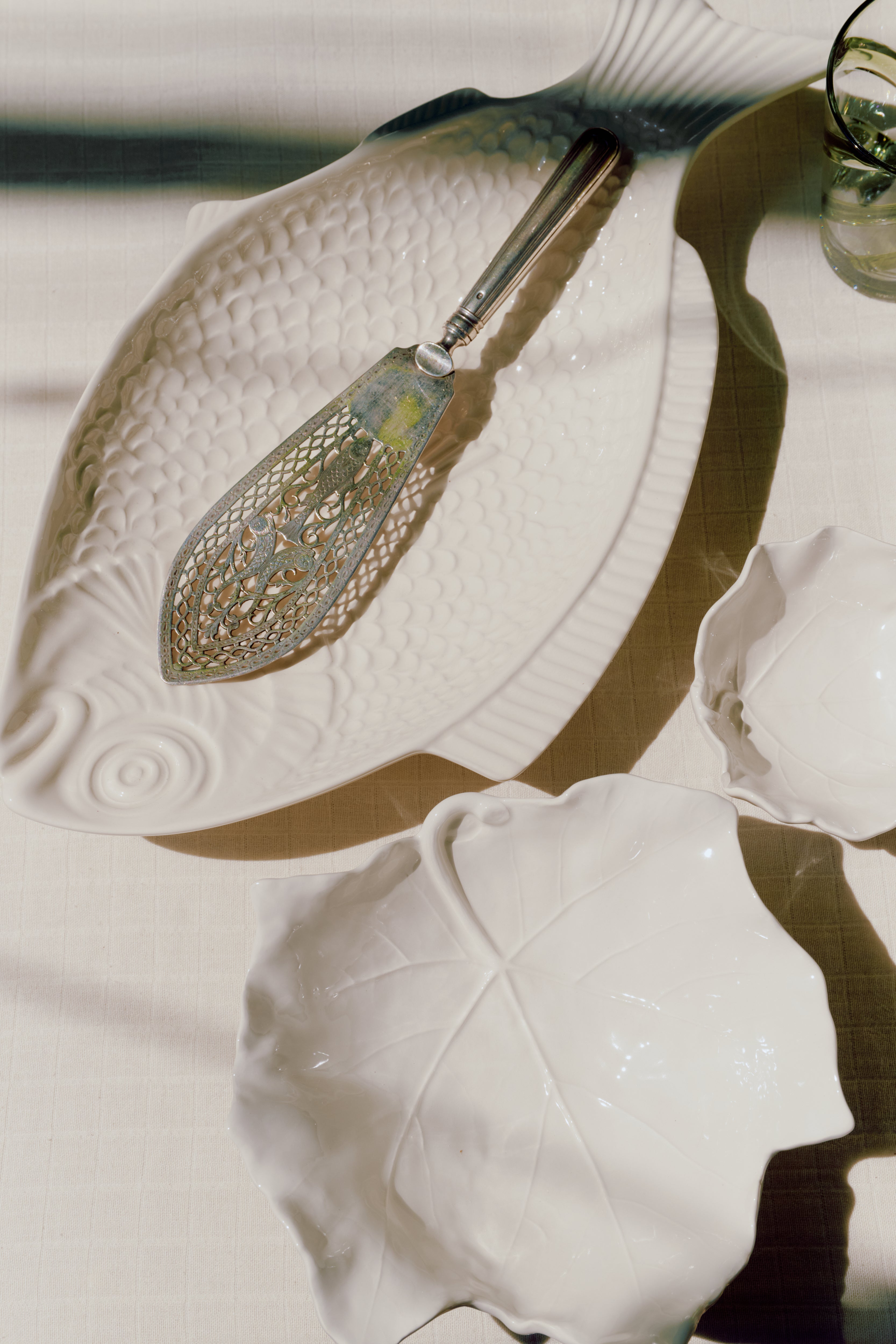 A metal fish server sits on a white ceramic fish-shaped dish, alongside two Leaf Serving Bowls, evoking the charm of an Italian countryside table on a light-colored tablecloth.
