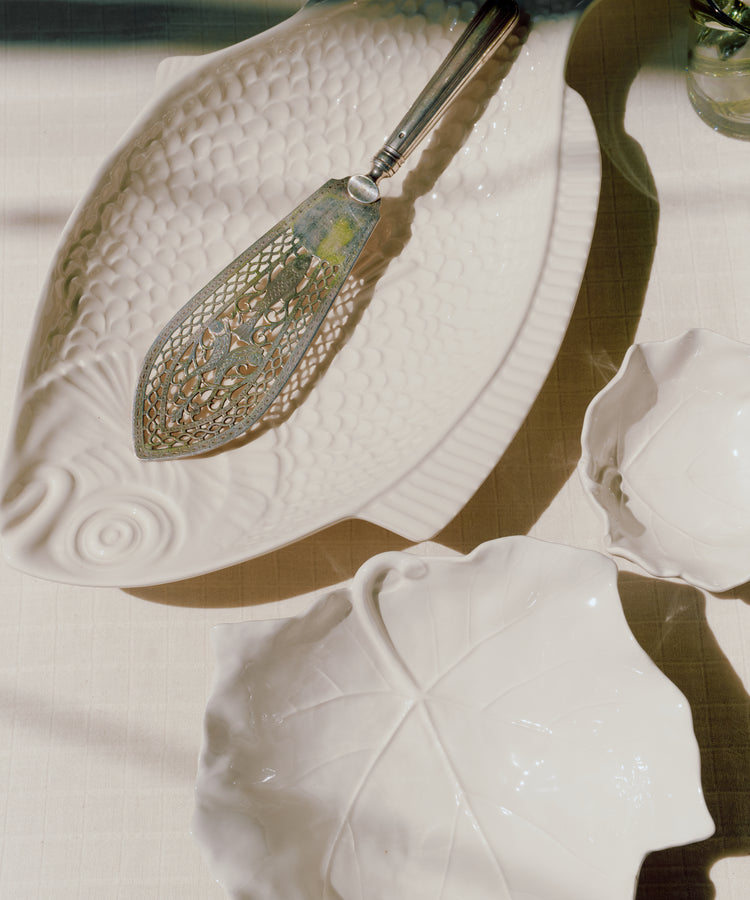 A metal fish server sits on a white ceramic fish-shaped dish, alongside two Leaf Serving Bowls, evoking the charm of an Italian countryside table on a light-colored tablecloth.
