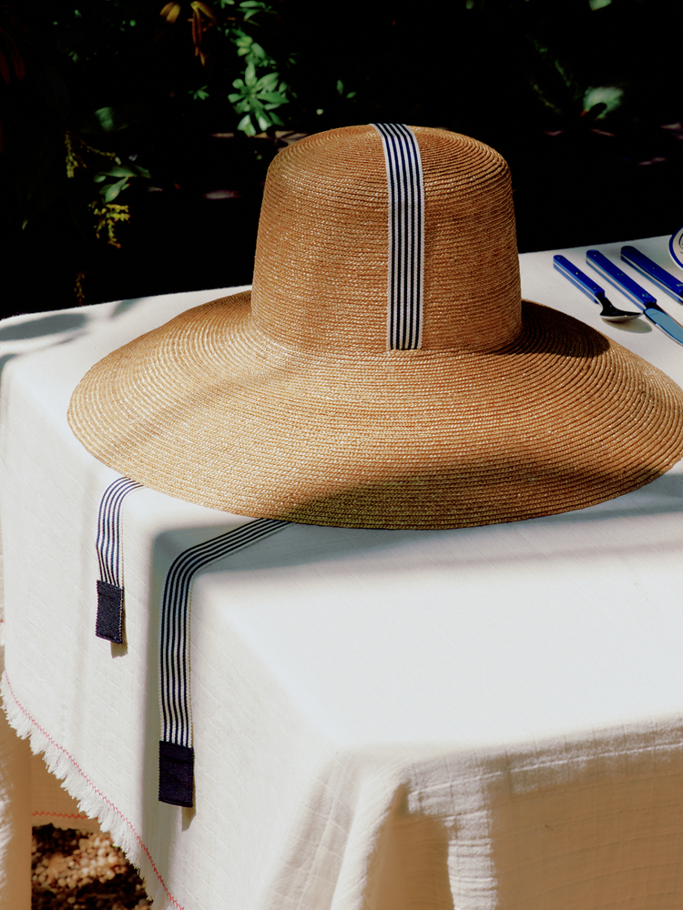 The Dahlia Hat, a natural straw style with a black-and-white striped ribbon, sits on a white tablecloth-covered table set with cutlery, its wide brim casting a soft floral silhouette in the afternoon light.