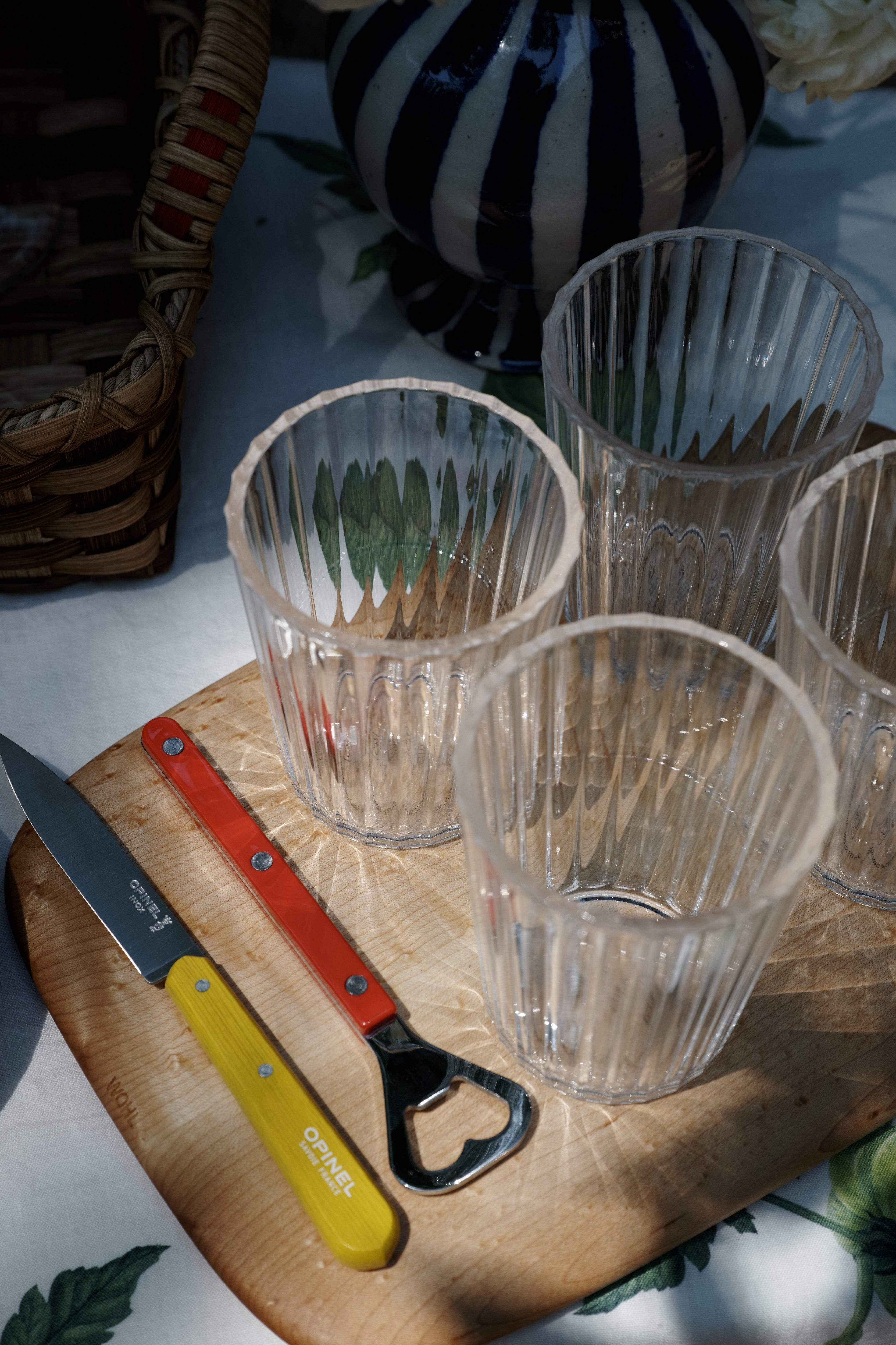 Four empty ribbed glasses, a yellow-handled bottle opener, and a red knife from the Classic Paring Knives, Set of 4 rest on a wooden board atop a table with a patterned tablecloth.