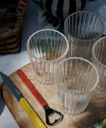 Four empty ribbed glasses, a yellow-handled bottle opener, and a red knife from the Classic Paring Knives, Set of 4 rest on a wooden board atop a table with a patterned tablecloth.