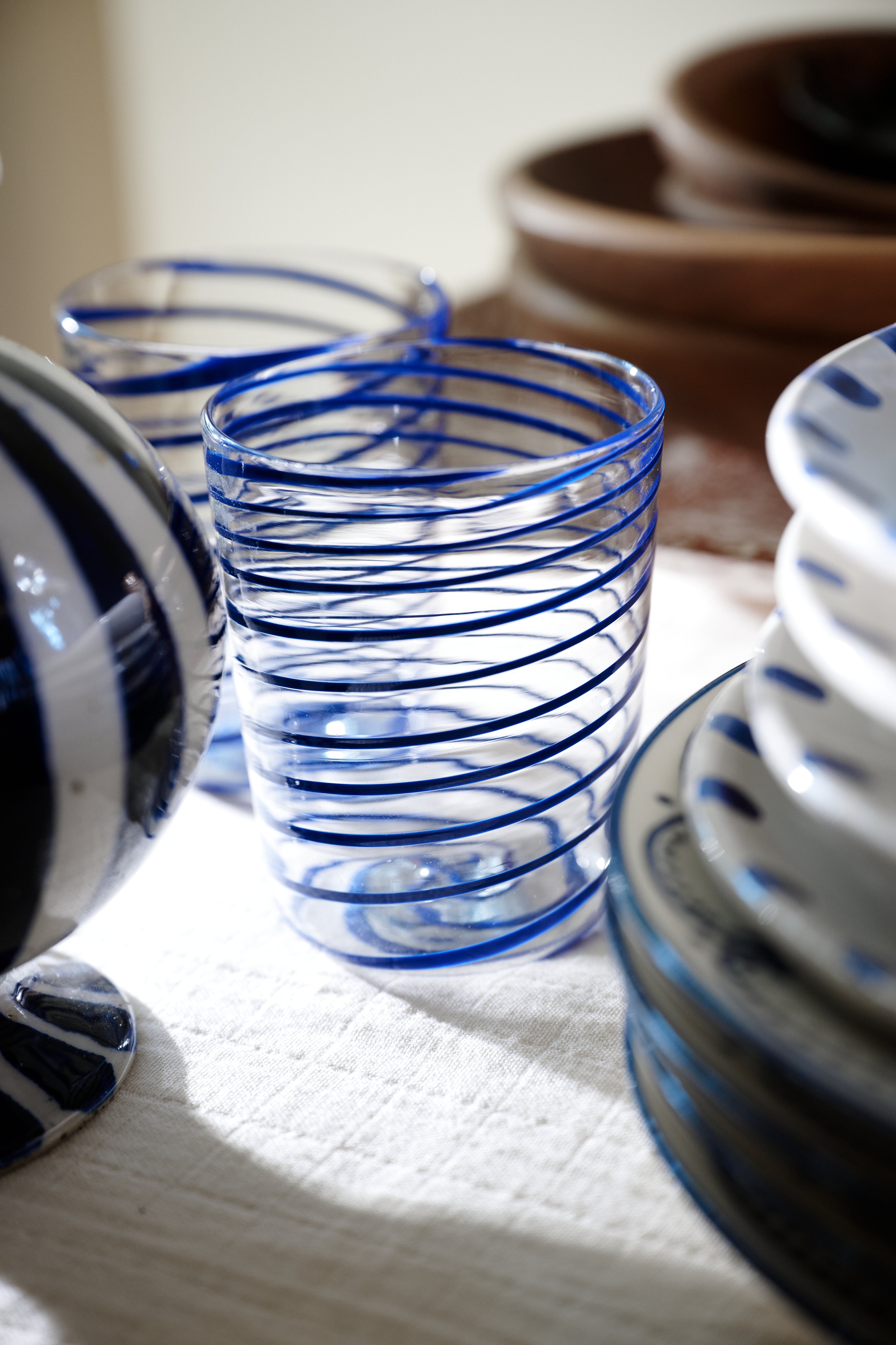 The Spirale Glass, Set of 2 features clear Murano-style spirals with cobalt blue patterns and adds Italian elegance when paired with stacked ceramic plates and brown bowls on a light cloth.