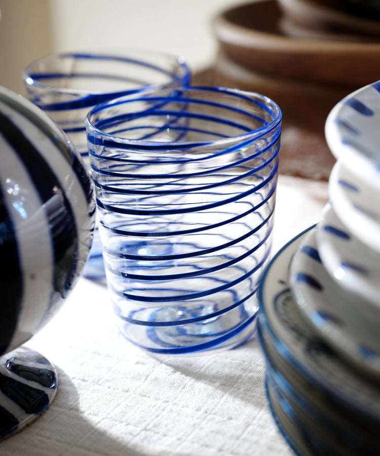 The Spirale Glass, Set of 2 features clear Murano-style spirals with cobalt blue patterns and adds Italian elegance when paired with stacked ceramic plates and brown bowls on a light cloth.