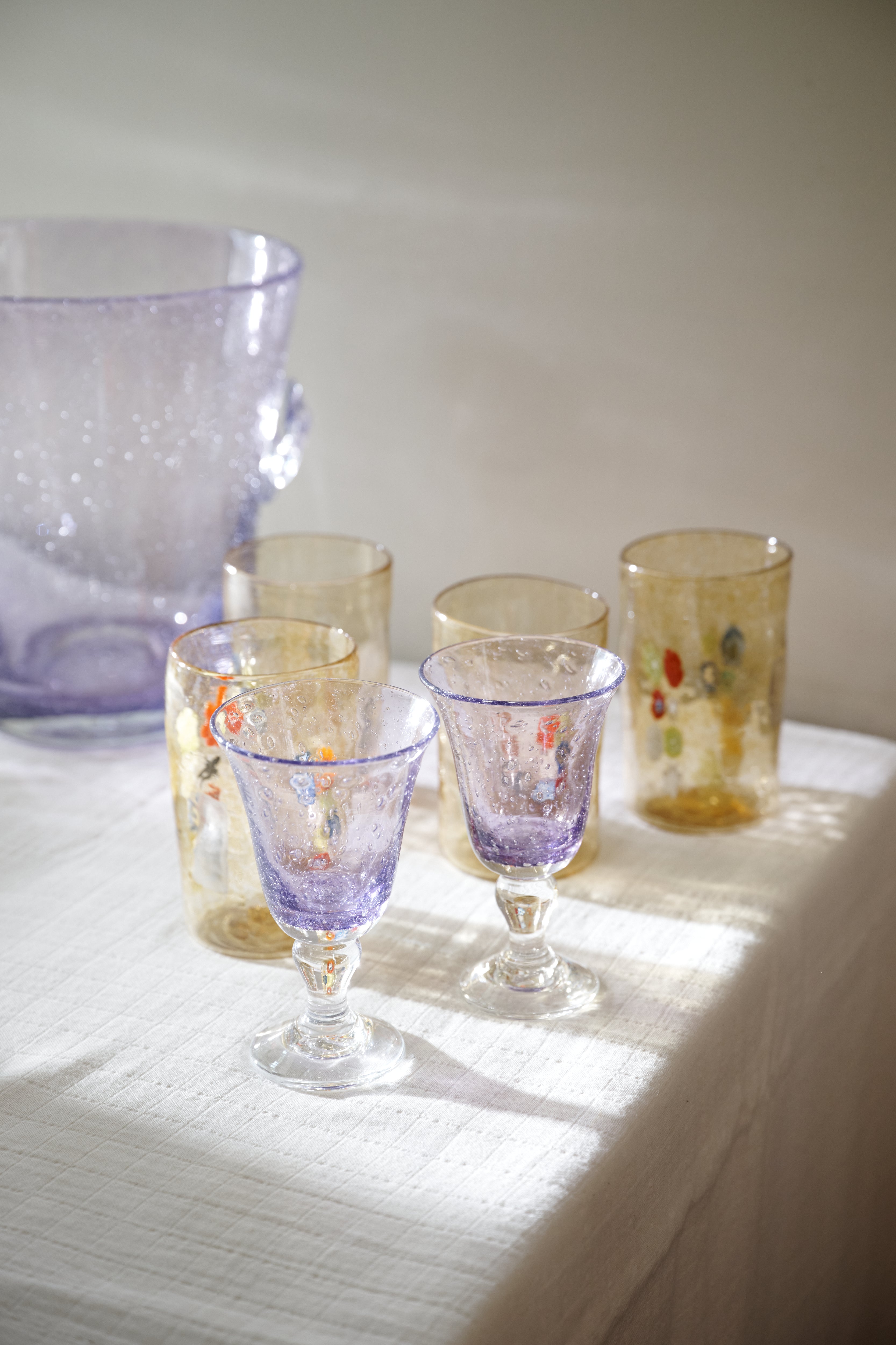A set of hand-blown glass tableware, including two purple goblets, two gold-toned tumblers with colorful floral details, and a large lavender pitcher, arranged on a white textured tablecloth in soft natural light.
