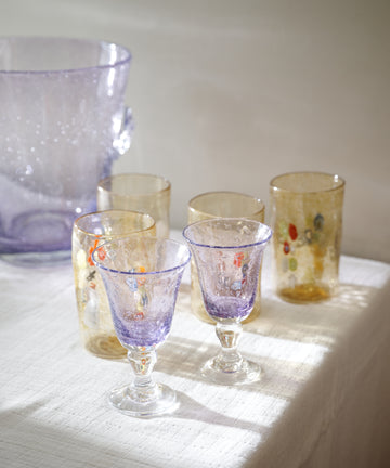 A set of hand-blown glass tableware, including two purple goblets, two gold-toned tumblers with colorful floral details, and a large lavender pitcher, arranged on a white textured tablecloth in soft natural light.