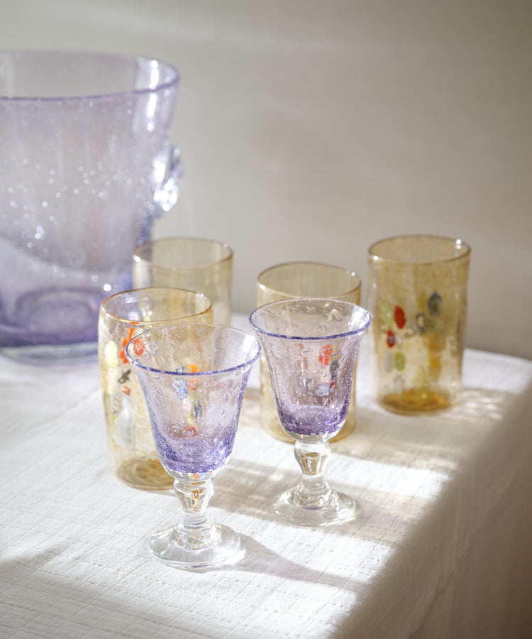 A set of hand-blown glass tableware, including two purple goblets, two gold-toned tumblers with colorful floral details, and a large lavender pitcher, arranged on a white textured tablecloth in soft natural light.