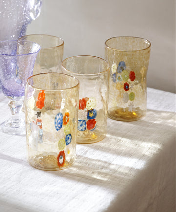 Two textured, handblown Muranesi Glasses featuring colorful floral patterns sit on a white tablecloth beside a purple goblet. Sunlight casts soft shadows, showcasing Venetian artisanal craftsmanship. Set includes 2 glasses.