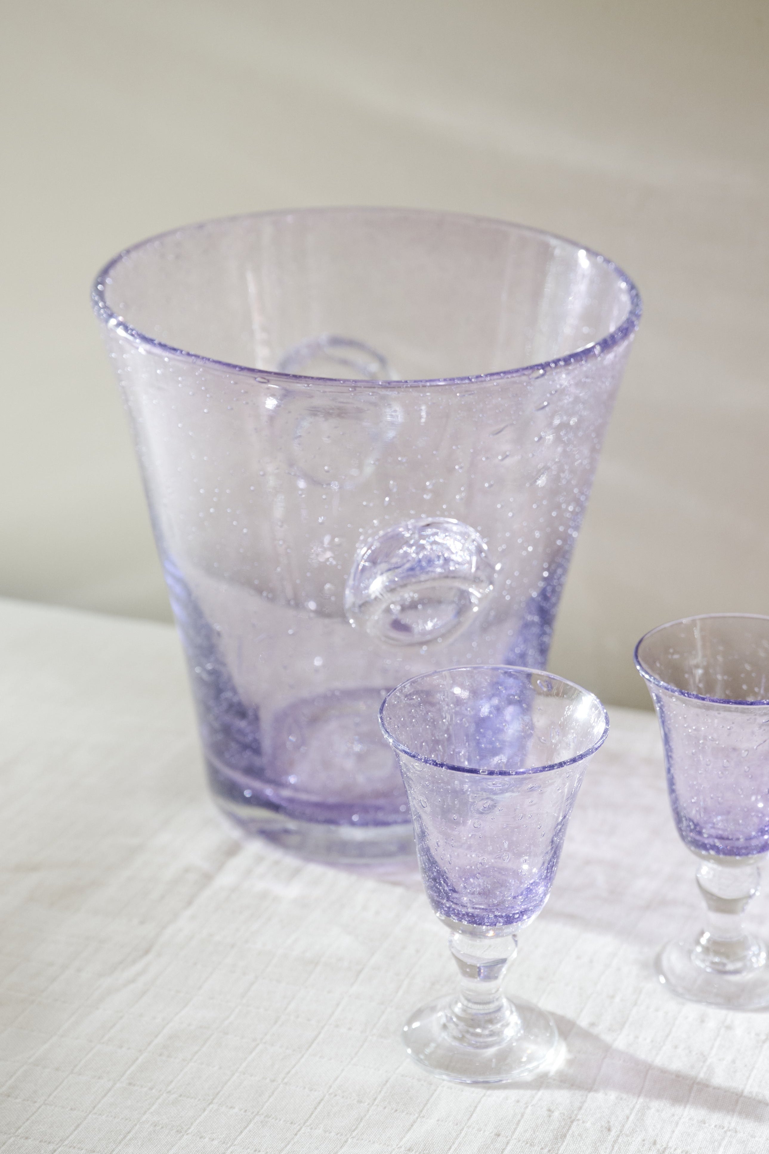 A set of three hand-blown glassware pieces in a light purple/lavender hue, including a large tapered pitcher and two small goblets, arranged on a white textured tablecloth.