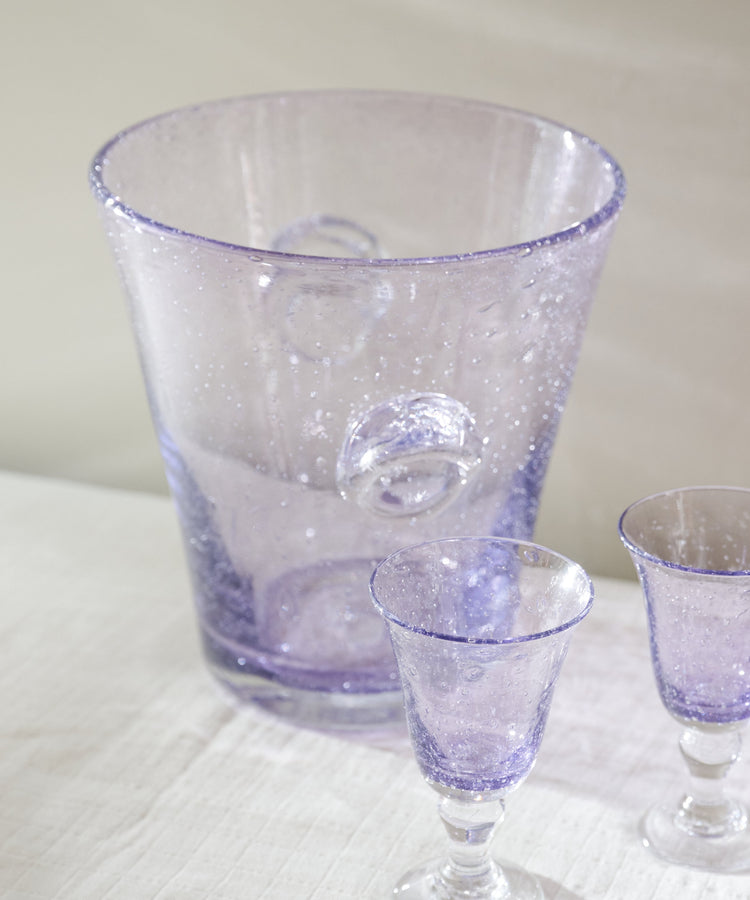 A set of three hand-blown glassware pieces in a light purple/lavender hue, including a large tapered pitcher and two small goblets, arranged on a white textured tablecloth.