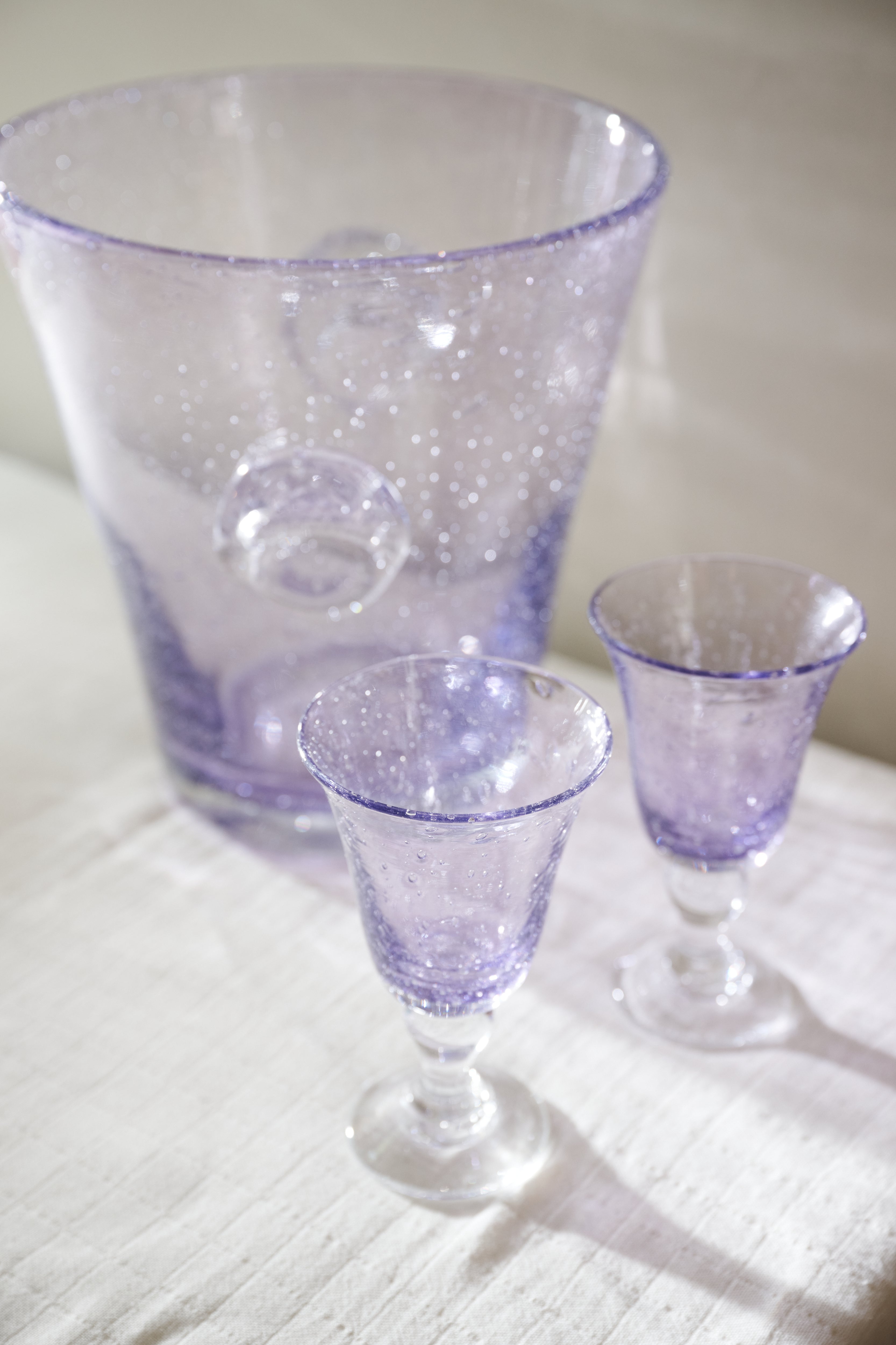 A purple bubble glass ice bucket and a set of two Lauren Wine Glasses are displayed on a white cloth surface.
