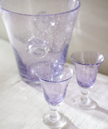 A purple bubble glass ice bucket and a set of two Lauren Wine Glasses are displayed on a white cloth surface.