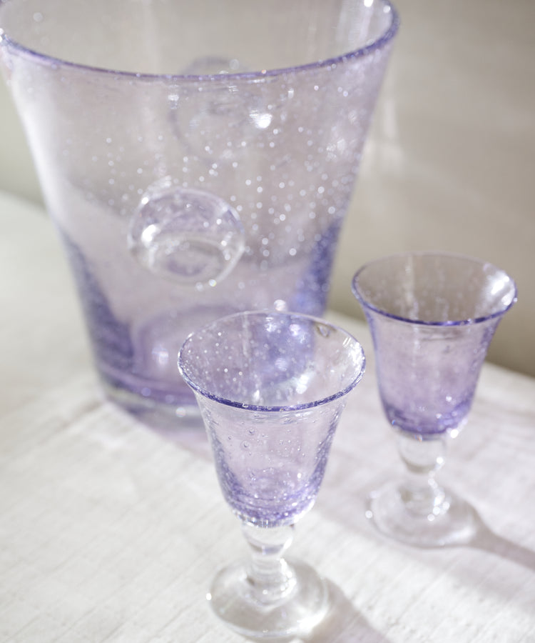 A purple bubble glass ice bucket and a set of two Lauren Wine Glasses are displayed on a white cloth surface.