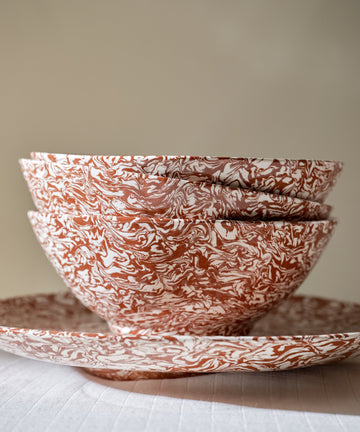 Three stacked Celine Salad Bowls, featuring a striking marbled red and white pattern, sit inside a matching plate on a white tablecloth against a neutral background.