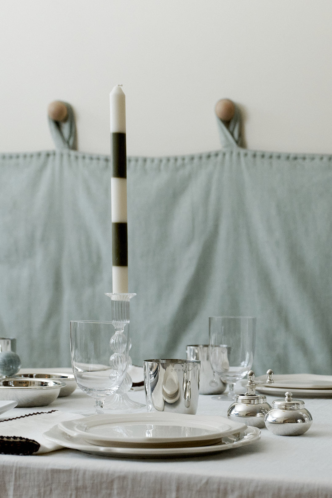A neatly set dining table with white plates, glassware, silver cups, a Hampstead Candle featuring black-and-white stripes, and silver containers on a white tablecloth.