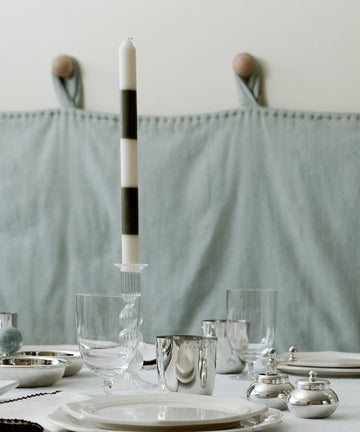 A neatly set dining table with white plates, glassware, silver cups, a Hampstead Candle featuring black-and-white stripes, and silver containers on a white tablecloth.