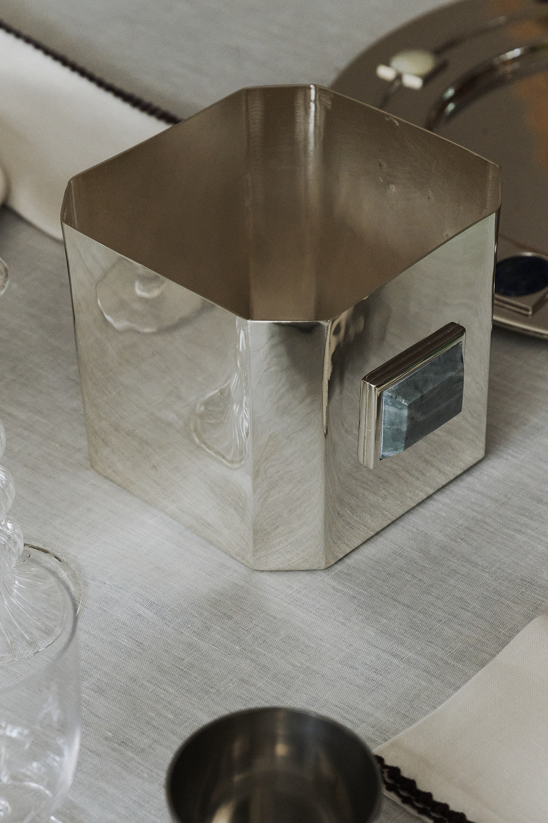 The Paros Square Vase, crafted from silver-plated brass with a geometric silhouette and a distinctive rectangular handle, rests on a white tablecloth beside glassware and a metal tray, evoking the minimalist elegance of a Brancusi vase.