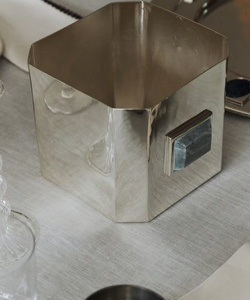 The Paros Square Vase, crafted from silver-plated brass with a geometric silhouette and a distinctive rectangular handle, rests on a white tablecloth beside glassware and a metal tray, evoking the minimalist elegance of a Brancusi vase.
