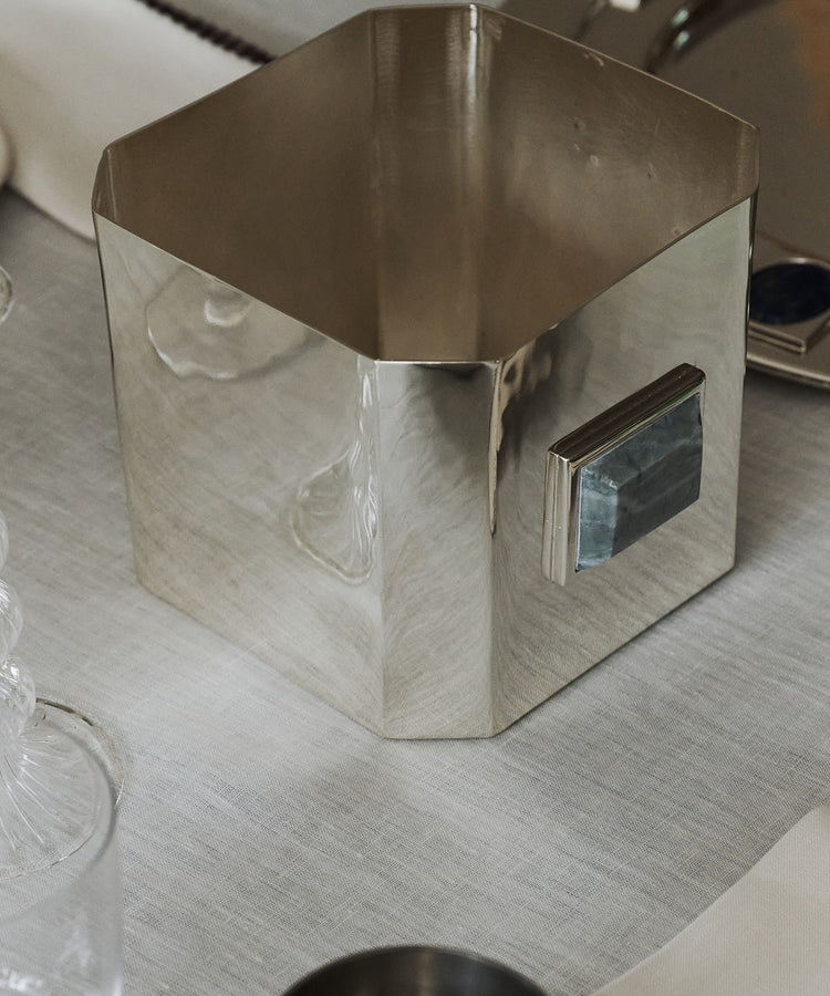 The Paros Square Vase, crafted from silver-plated brass with a geometric silhouette and a distinctive rectangular handle, rests on a white tablecloth beside glassware and a metal tray, evoking the minimalist elegance of a Brancusi vase.