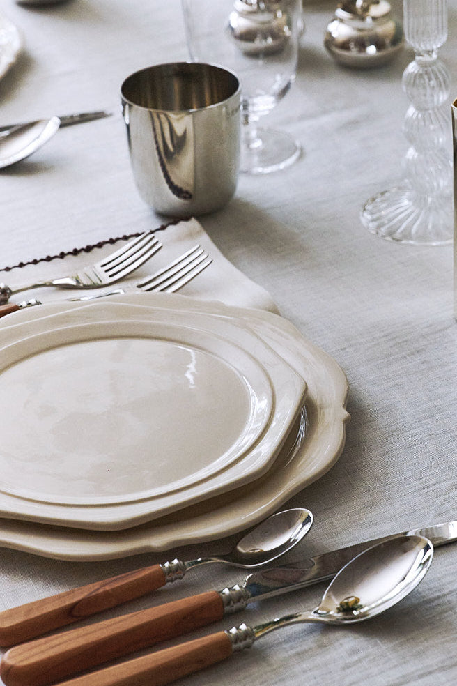 A styled dining table setting with stacked white plates, silver-toned cutlery with light wood handles, clear glassware, and a reflective silver container holding yellow lemons. The table is covered with a light gray linen tablecloth.