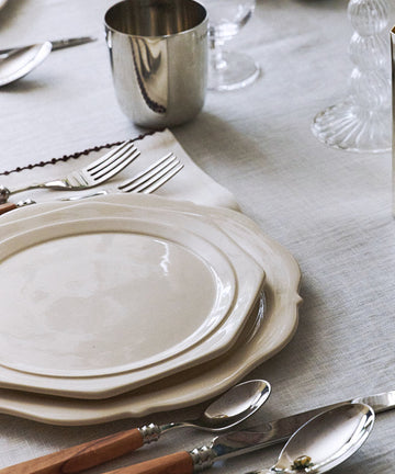 A styled dining table setting with stacked white plates, silver-toned cutlery with light wood handles, clear glassware, and a reflective silver container holding yellow lemons. The table is covered with a light gray linen tablecloth.