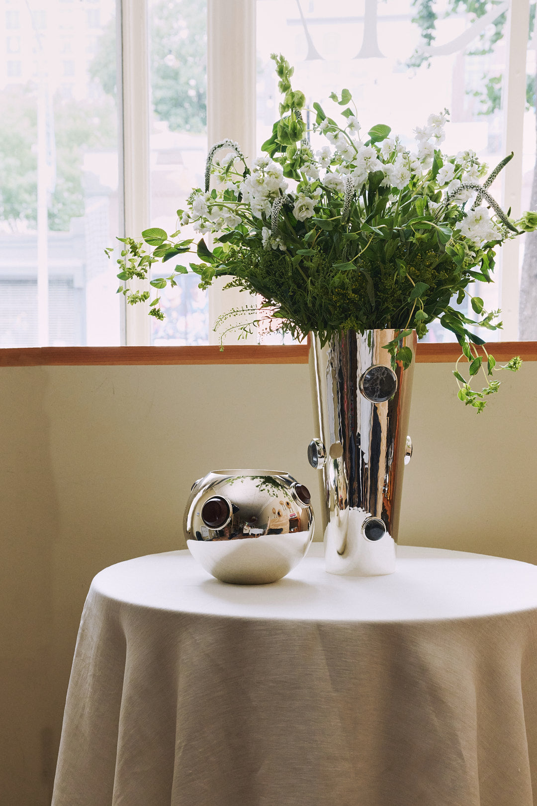 A round table with a white tablecloth holds a shiny handmade Paros Cone Vase filled with white flowers and greenery, next to a small silver decorative orb.