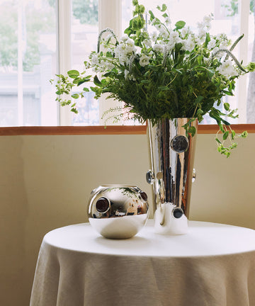 A round table with a white tablecloth holds a shiny handmade Paros Cone Vase filled with white flowers and greenery, next to a small silver decorative orb.