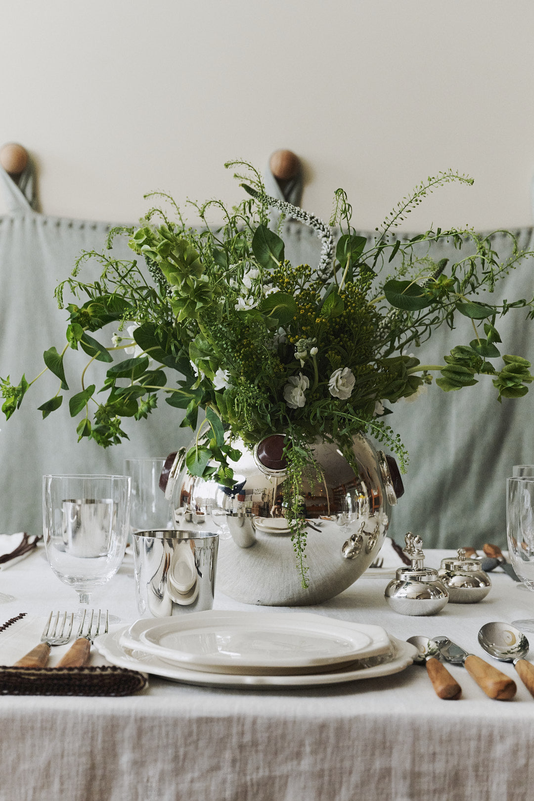 The Paros Round Vase with lush green foliage serves as the centerpiece on a white tablecloth, surrounded by plates, glasses, cutlery, and napkins prepared for dining. The sophisticated tablescape is further elevated by accents of marble decor that enhance the refined atmosphere.