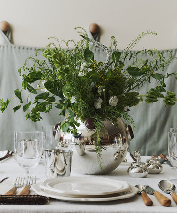 The Paros Round Vase with lush green foliage serves as the centerpiece on a white tablecloth, surrounded by plates, glasses, cutlery, and napkins prepared for dining. The sophisticated tablescape is further elevated by accents of marble decor that enhance the refined atmosphere.