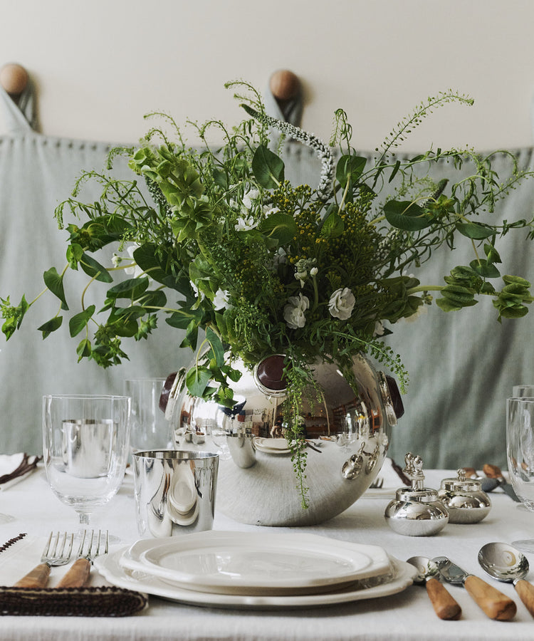 The Paros Round Vase with lush green foliage serves as the centerpiece on a white tablecloth, surrounded by plates, glasses, cutlery, and napkins prepared for dining. The sophisticated tablescape is further elevated by accents of marble decor that enhance the refined atmosphere.